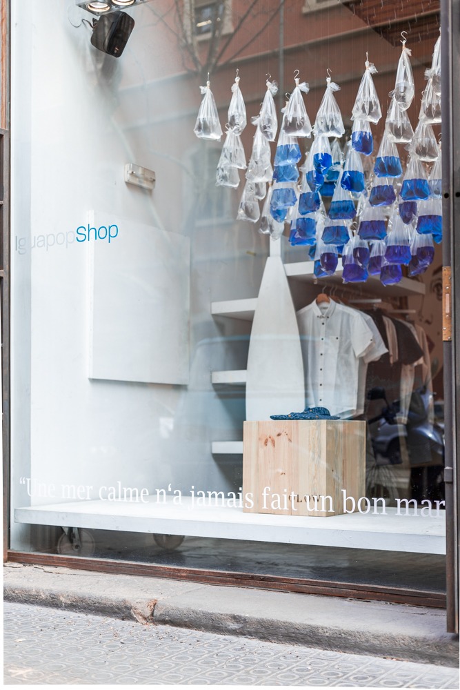 For this Spring/Summer season, Iguapop in Barcelona has a new shop window starring OLOW! Created by a group of students from the Elisava School of Design in Barcelona, who won a contest to conceive their Liquid Modernity concept, the window was inspired by a saying found in the catalogue of our Marée Basse collection: "Une mer calme n'a jamais fait un bon marin", or "Smooth seas never made a skilled sailor". This concept portrays a sailor who tries to navigate through society, trying to keep his principles afloat despite the storm. To reinterpret this society, the students were inspired by Zygmunt Bauman’s book Liquid Modernity, which introduced this idea of liquid modernity: a society represented by a man faced with uncertainty and ambivalent feelings, a nomad, a man with no traditional patterns but self-chosen ones, with an emphasis on change and an immersion into the fast-paced consumerism of our time. The changing and tormented seas are thus a metaphor for this society and its volume economy. On the contrary, OLOW, the sailor, portrays "slow fashion", transmitting more traditional and honest values, prioritising to quality over quantity. This idea shapes the showcase in the Iguapop Shop, which a few years ago was also a contemporary art gallery and still maintains that essence. With the proposed design, the shop window forms part of the shop’s interior, creating a specific space for the brand within its commercial space. We at OLOW are incredibly pleased and flattered to be the window’s guest of honour, so we decided to interview the talented designers to get to know them more and better understand their concept. We also asked Eva Lluch, Iguapop’s owner, a few questions about her shop and her desire to feature art prominently inside it. Hello everyone! You are students at the Elisava School of Design in Barcelona; could you tell us about yourselves and how you feel upon designing this incredibly creative and beautiful window?  Hello! For us, the success of our project is thanks to the team members. We are a mix of personalities and all have our own distinct abilities. The fact that we come from such varied backgrounds and are of different ages was definitely key: Anayansi Diaz is a 24-year-old industrial designer from Mexico DF, Marta Meléndez Rújula is also 24 and an industrial design engineer from Zaragoza in Spain, Joaquin Acevedo Cordon is a 31-year-old industrial designer from Chile and finally, Micaela Seresini Fernandez is a 28-year-old architect from Mar del Plata in Argentina. Having diverse understandings and multiple points of view on one same brief and concept gave us infinite options and ideas to explore! On the other hand, we think work relies on the exploration of how we construct our identities and how we all have a fascination for speculating over the “FUTURE”. What was your starting point to the concept? What was your main inspiration? From a very personal point of view, the catalogue’s aesthetic with its very evident marine theme was the trigger. The proverb “Une mer calme n’a jamais fait un bon marin” ("Smooth seas never made a skilled sailor") was definitely an inspiration. Every single thing we thought about orbited around the idea of the sea and the various difficulties a sailor could face. This visual imagery was fused with Zygmunt Bauman’s “Liquid modernity” theory that portrays the characteristics of a postmodern society and its constant search and thirst for change.   How long did the creative process take?  It took about three weeks. We brainstormed ideas for about a week, and once we had the idea and concept it took us the remaining two weeks to project the idea and determine whether it was actually doable in terms of cost and materialisation.   What do you want to say through this installation? The main message, besides obviously talking about OLOW as a brand, was to make a statement against our current lifestyle and society. The upper structure represents our consumer society and the liquid modernity phenomenon: we are constantly changing and eager to always have the newest things; while the wooden cube calls to mind the timeless aesthetics of well-made products that last and are fashionable no matter the time and context.   Technically, how did you create it? Once we had the idea and concept, everything came quite easily. The upper structure was a simple but very exhaustive exercise in parametric design. A sea wave was the core of this installation. We drew this trigonometric function curve (F(x)=cos x) to determine the lateral view and overlap it with a simple quadratic function (F(x)=x2). After this was settled, we had to work with the individual dimensions of the fish bags to determine the space between each bag on the x, y, z plans. That was the most complicated… along with the logistics of how to execute it in real life. The cube that represents OLOW and its values is a beautiful piece of woodcraft.   What did you learn from this experience? That different is always good, that change is imminent but does not imply creating just to create. And that making things correctly and with passion is the key to success. Eva Lluch is the manager of Iguapop in Barcelona. The shop now has an OLOW-inspired window created by a group of talented, young designers. We asked her a few questions concerning this great project. Your shop used to also feature an art gallery; are you passionate about art and is it your wish to continue to promote all kinds of art forms? Is it important for you to give these kinds of opportunities to young designers? Yes, of course, our store is linked to the culture of art and design. We have organised several art exhibitions and asked different artists to create textile caps for us. We like to galvanize our space with art and design-related events. Did you get involved in the creative process or did you give free-range to the designers? I was not involved in this creative process; it was a contest between several students of the ELISAVA school, where 8 projects competed to present OLOW’s new collection in Iguapop Shop’s window. I chose the winning project that would be exhibited.
